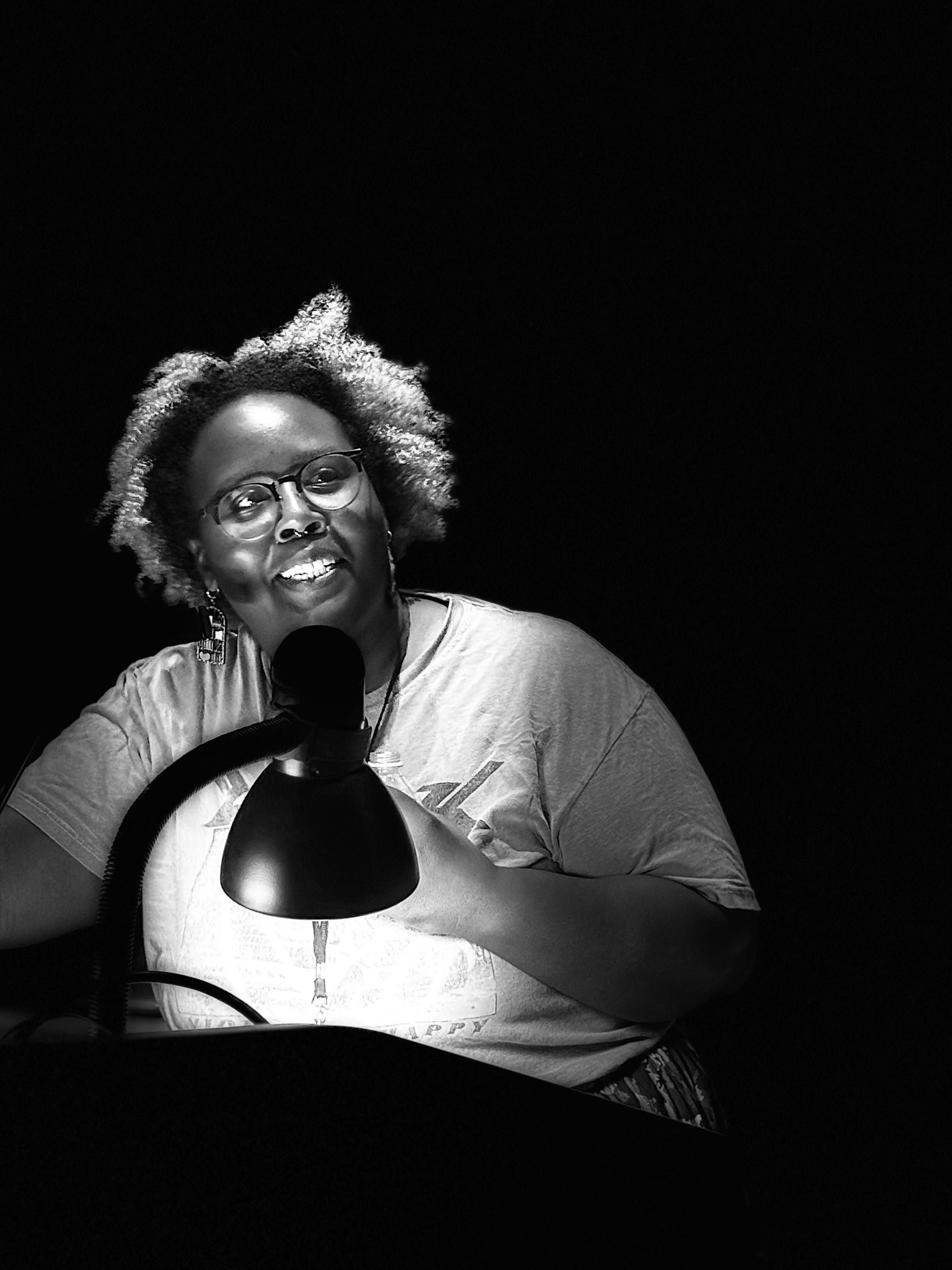 A smiling woman with curly hair speaks passionately at a podium, illuminated by a desk lamp, against a dark background.